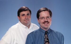Two men pose for a studio portrait against a blue backdrop, one in a white shirt and the other in a dress shirt and tie.