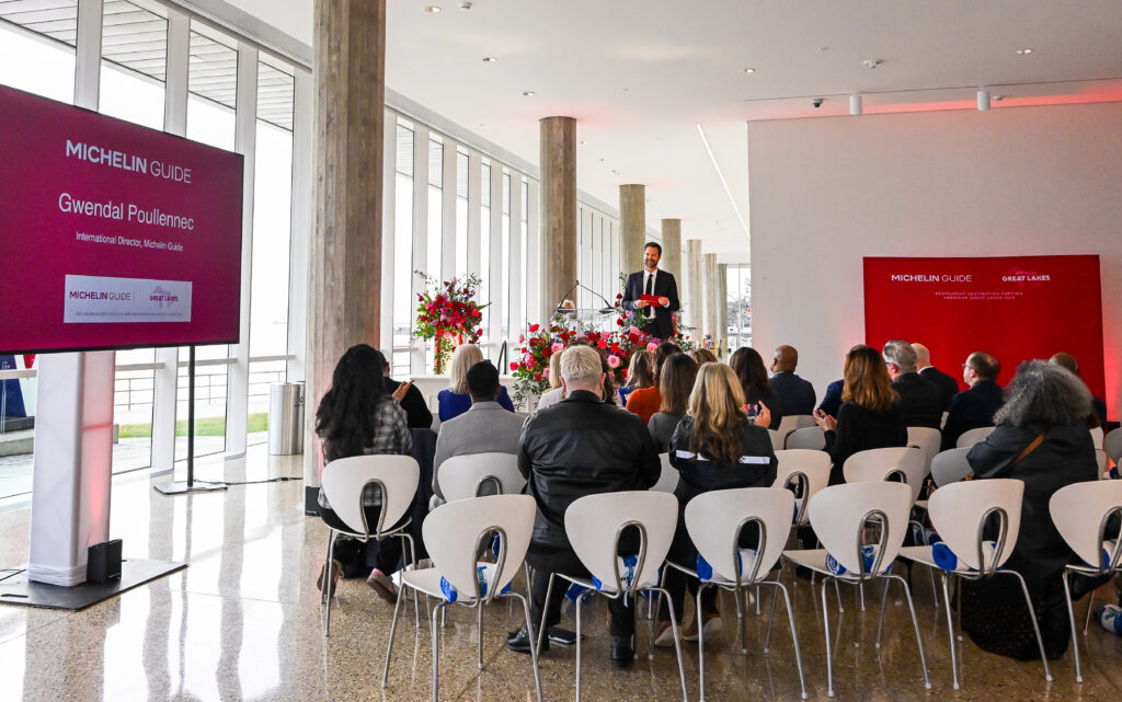 A speaker stands at a podium addressing a seated audience at a Michelin Guide event, with large screens displaying “Michelin Guide” and “Great Lakes” branding.
