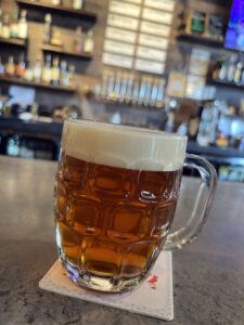 A dimpled glass mug of amber beer with a thick foamy head sits on a bar counter, with taps, bottles, and a menu board softly blurred in the background.