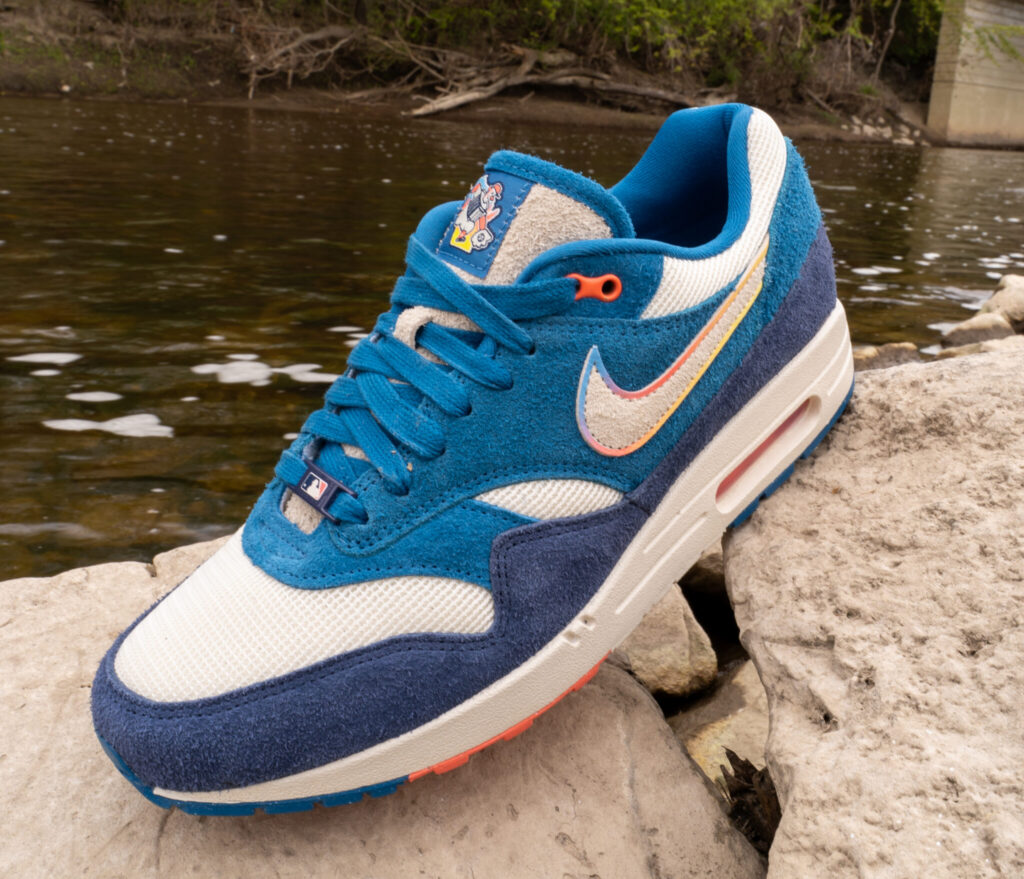 A blue, white, and navy Nike sneaker with a multicolor swoosh sits on a rock beside a shallow river, with trees and a bridge in the background.