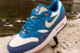 A blue, white, and navy Nike sneaker with a multicolor swoosh sits on a rock beside a shallow river, with trees and a bridge in the background.