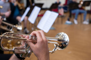Close-up of a person playing a silver trumpet during a band rehearsal, with blurred musicians and sheet music stands visible in the background.