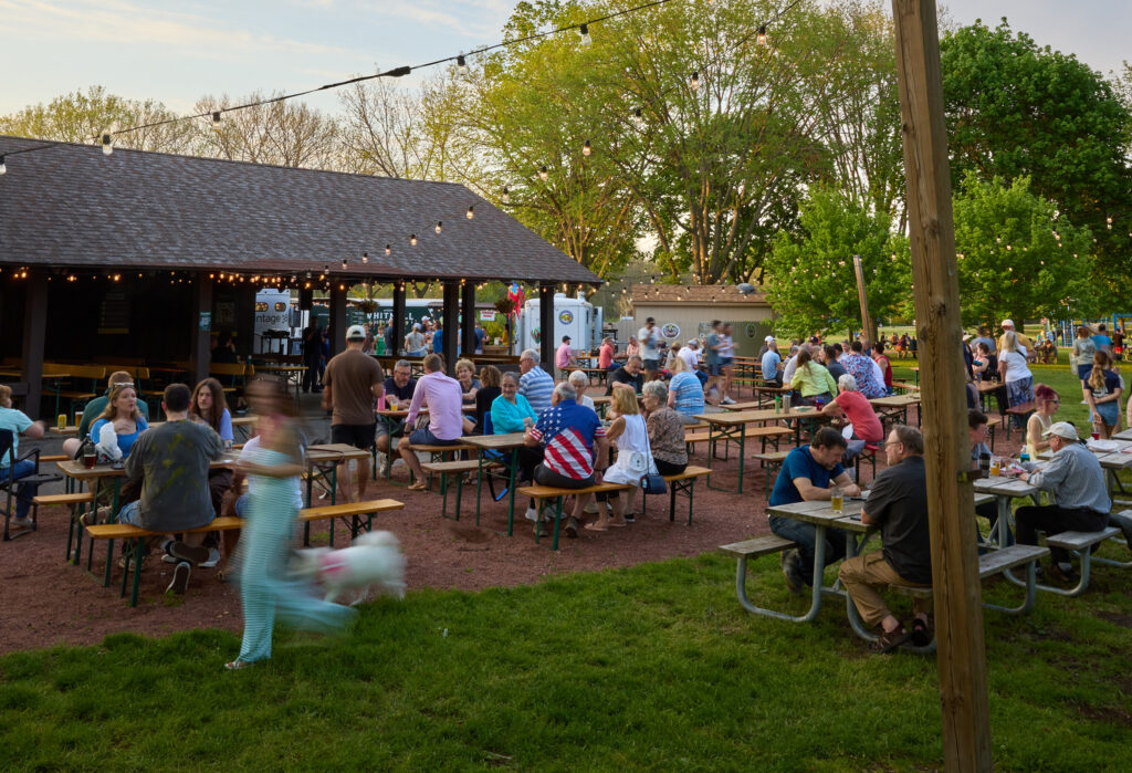 People gather at picnic tables in an outdoor beer garden at dusk, with string lights overhead, a pavilion and food trucks in the background, and families and groups socializing on the lawn.