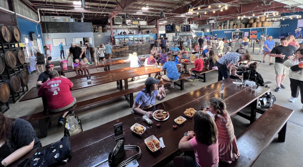 A large brewery taproom with long communal tables is filled with families and groups eating, drinking, and socializing, with beer barrels, string lights, and a bar area visible throughout the industrial-style space.