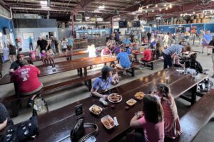 A large brewery taproom with long communal tables is filled with families and groups eating, drinking, and socializing, with beer barrels, string lights, and a bar area visible throughout the industrial-style space.