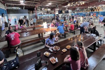 A large brewery taproom with long communal tables is filled with families and groups eating, drinking, and socializing, with beer barrels, string lights, and a bar area visible throughout the industrial-style space.