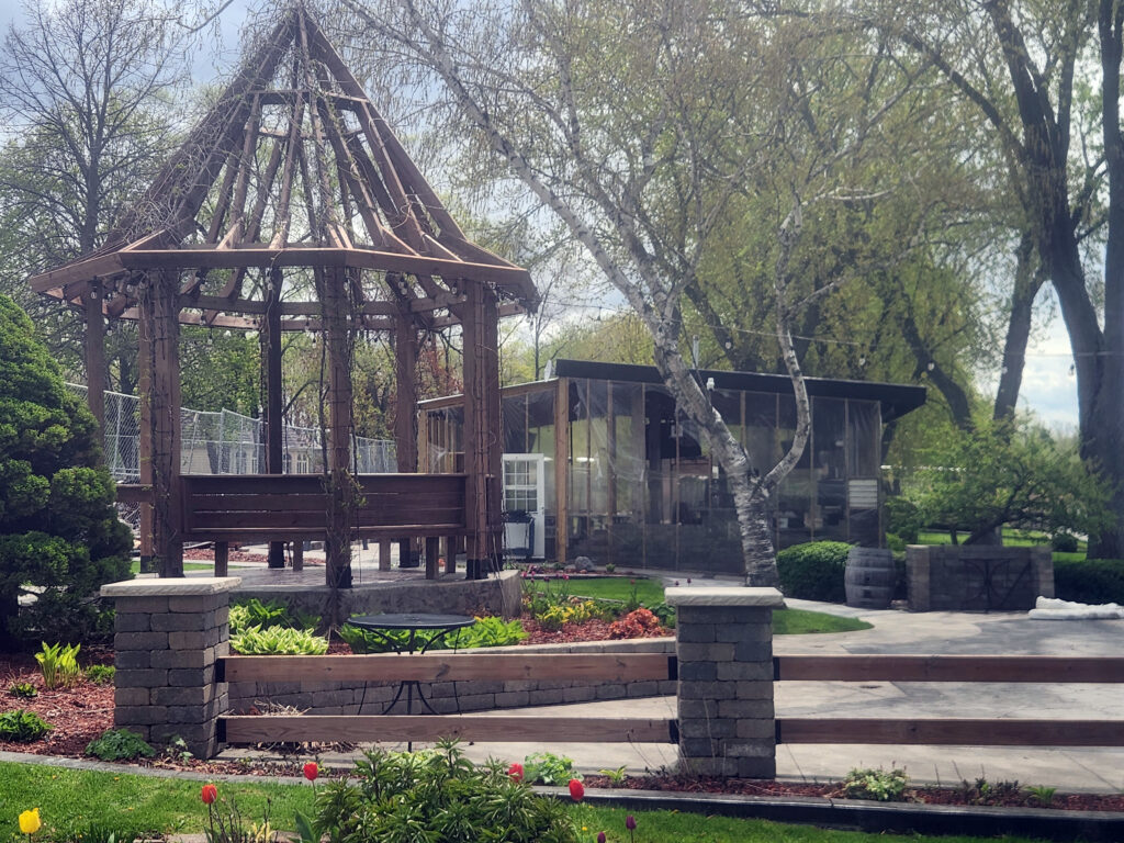 A wooden gazebo with a peaked, open frame stands in a landscaped garden among trees, a small building with plastic sheeting in front of a cyclone fence.