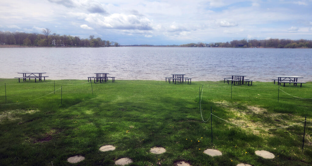 Picnic tables sit spaced across a grassy lakeshore overlooking a wide, calm lake under a partly cloudy sky.