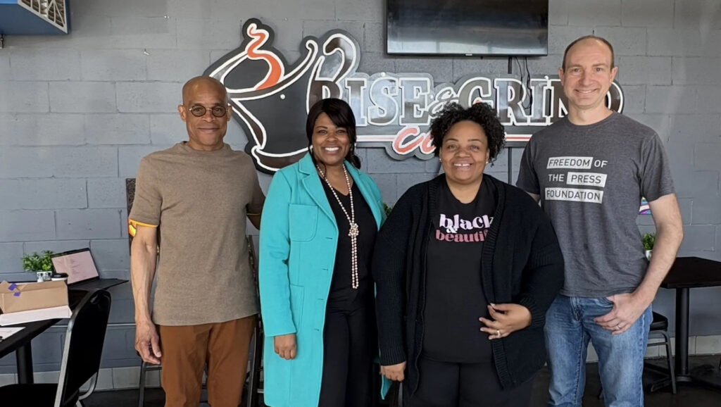 Four people stand together inside a cafe in front of a wall logo reading “Rise & Grind,” smiling for a group photo, with one person wearing a shirt that reads “Freedom of the Press Foundation.”