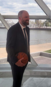 A man in a dark suit smiles while holding a basketball inside a glass-walled space overlooking the waterfront.