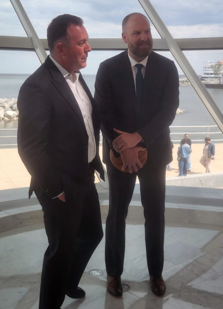 Two men in suits stand inside a lakeside atrium, one holding a basketball, with water and a pier visible through large windows behind them.