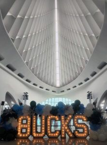 Large illuminated letters spelling “BUCKS” sit beneath a high, ribbed white ceiling inside a modern atrium, decorated with green, blue, and white balloons.