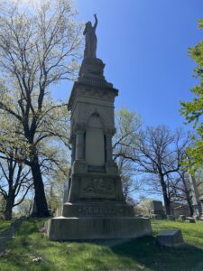 A tall stone monument topped with a statue stands in a cemetery beneath a clear blue sky, with the name “Schlitz” carved into the base and large trees surrounding the gravesite.