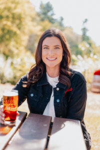 A woman in a black jacket sits outdoors holding a glass of amber-colored beer.