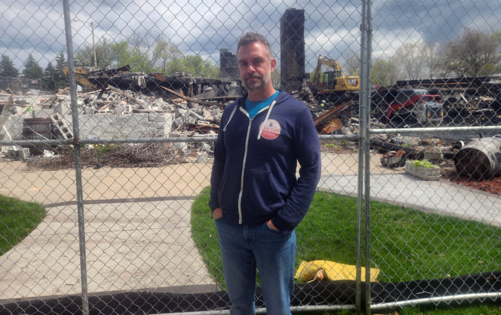 A man wearing a navy zip-up hoodie with a circular logo, a blue T-shirt, and jeans stands in front of a chain-link fence with the charred remains of a demolished building and heavy equipment behind him.
