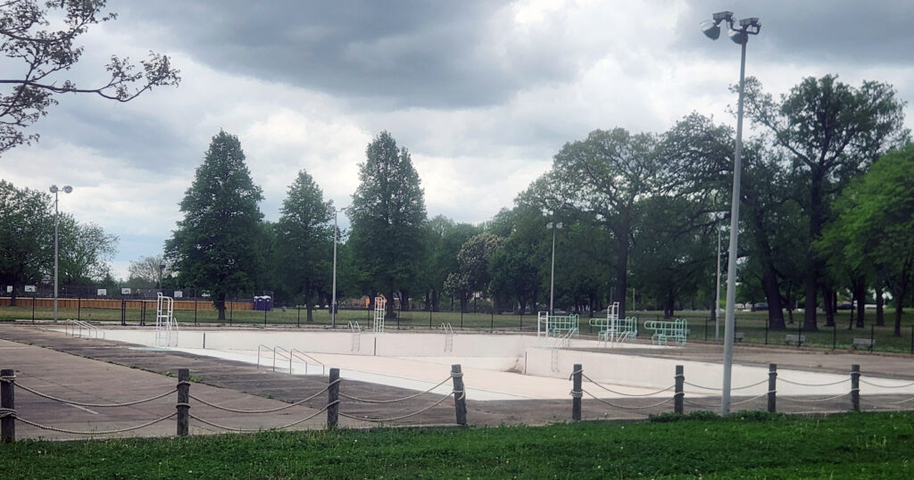 A drained public swimming pool with ladders and diving structures stands empty beneath dark clouds, surrounded by trees and park grounds.