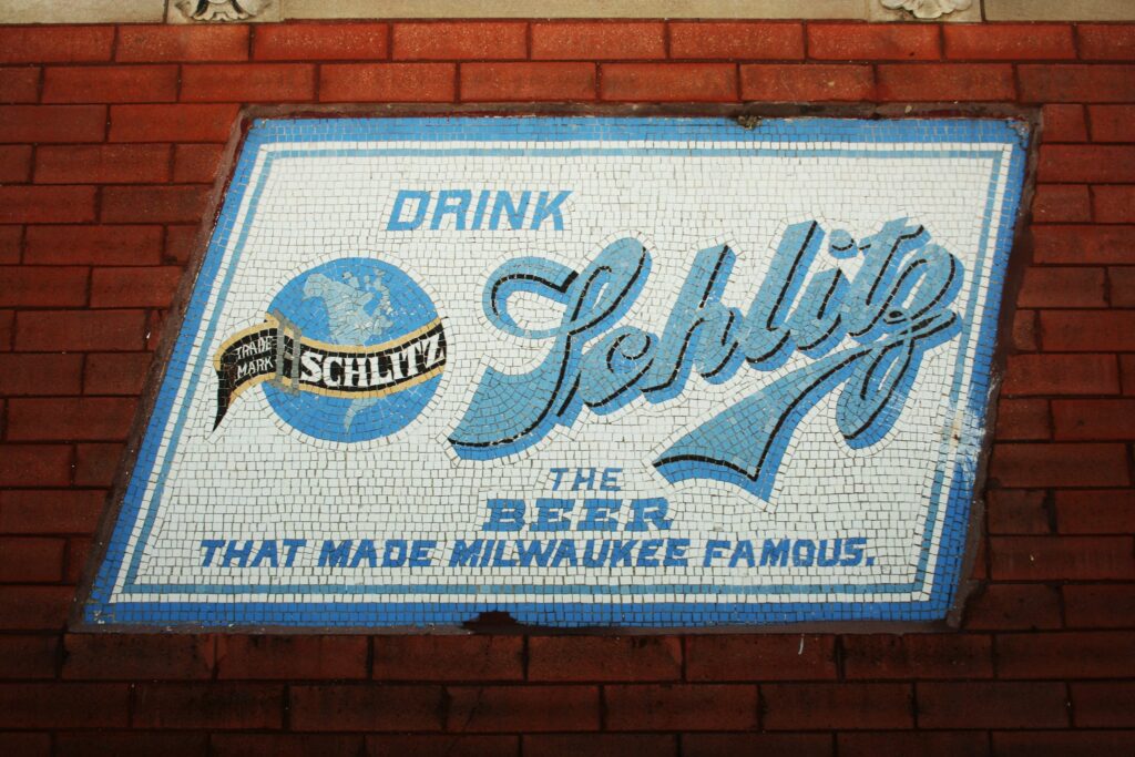 A vintage Schlitz beer mosaic sign set into a red brick wall reads “Drink Schlitz, the beer that made Milwaukee famous,” with blue and white tile lettering and the brand’s globe logo.