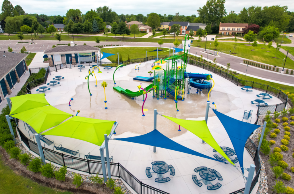 Aerial rendering of a modern splash pad and water playground with slides, spray features, shaded seating areas, and surrounding landscaping in a suburban park setting.