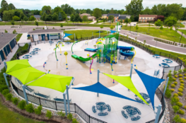 Aerial rendering of a modern splash pad and water playground with slides, spray features, shaded seating areas, and surrounding landscaping in a suburban park setting.