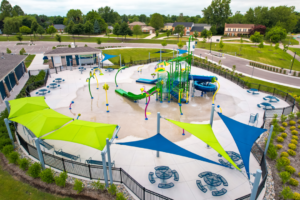 Aerial rendering of a modern splash pad and water playground with slides, spray features, shaded seating areas, and surrounding landscaping in a suburban park setting.