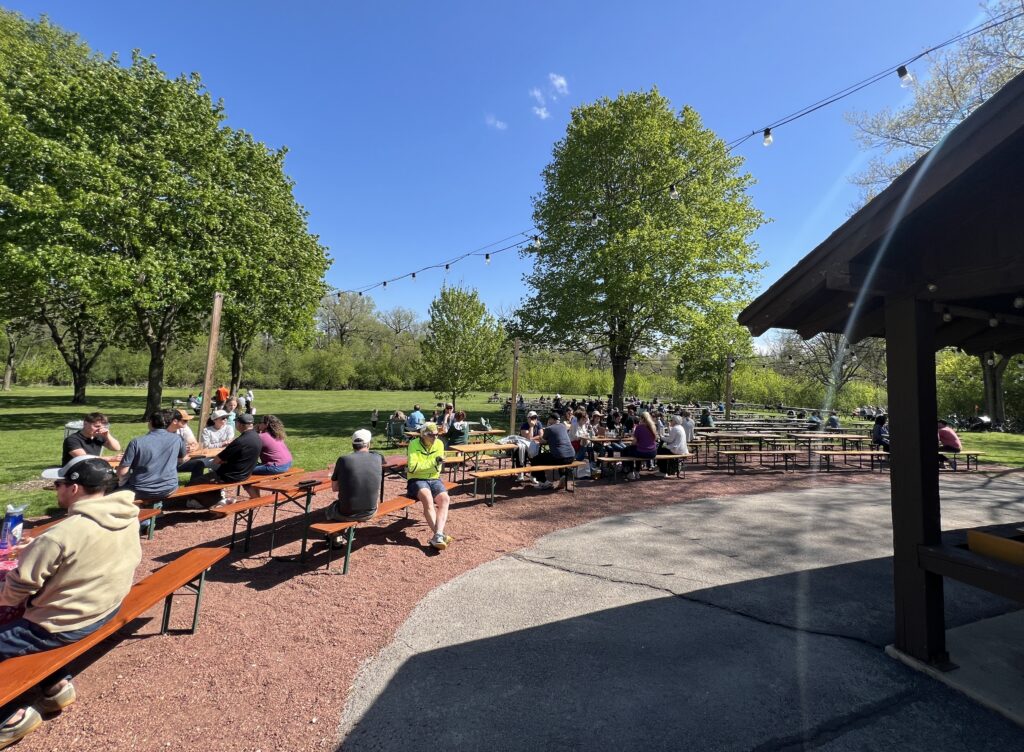 People sit at long picnic tables in a spacious outdoor beer garden surrounded by trees and open green space, with string lights hanging overhead on a sunny day.