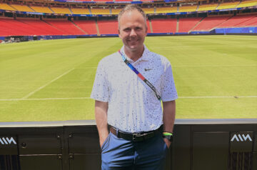 A man in a white patterned polo shirt and blue shorts stands on the sideline of a soccer stadium, with a scoreboard displaying “FIFA World Cup 2026” above rows of red and yellow seats and a green playing field behind him.