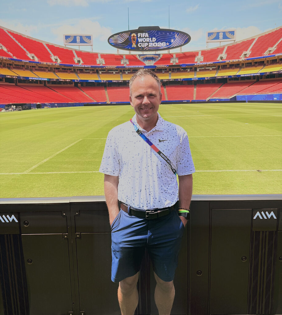 A man in a white patterned polo shirt and blue shorts stands on the sideline of a soccer stadium, with a scoreboard displaying “FIFA World Cup 2026” above rows of red and yellow seats and a green playing field behind him.