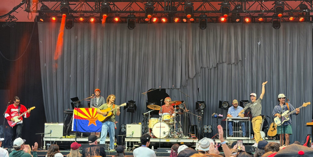 A band performs on an outdoor festival stage as concertgoers gather in front of the crowd barrier, with an Arizona state flag displayed near the musicians.
