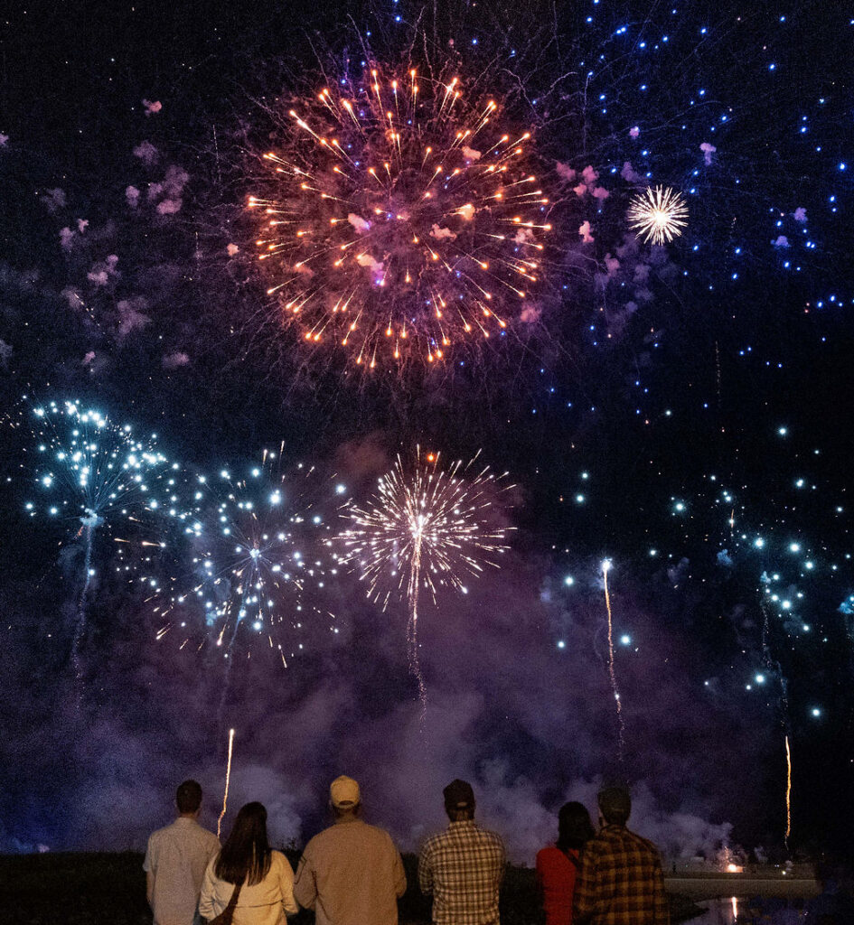 People watch a fireworks display as colorful bursts of red, blue, and white light fill the night sky above a waterfront.