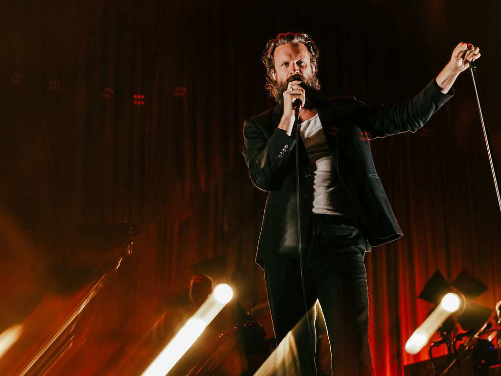 A bearded singer performs onstage under dramatic lighting, holding a microphone stand in front of a backdrop of red curtains.
