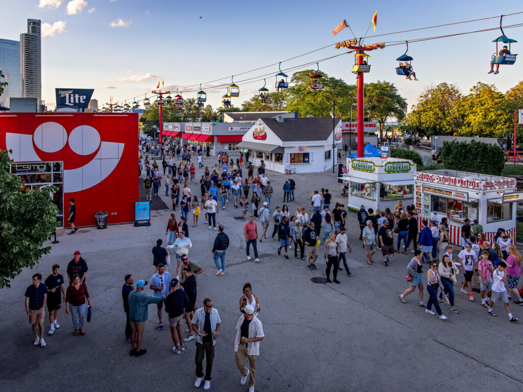 Crowds stroll through the Summerfest grounds past food stands, festival buildings, and an overhead Skyglider ride on a summer evening.