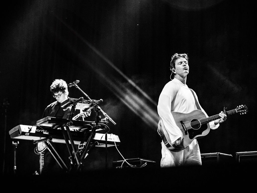 A guitarist dressed in white and keyboardist perform onstage under spotlights during a concert, captured in black and white.