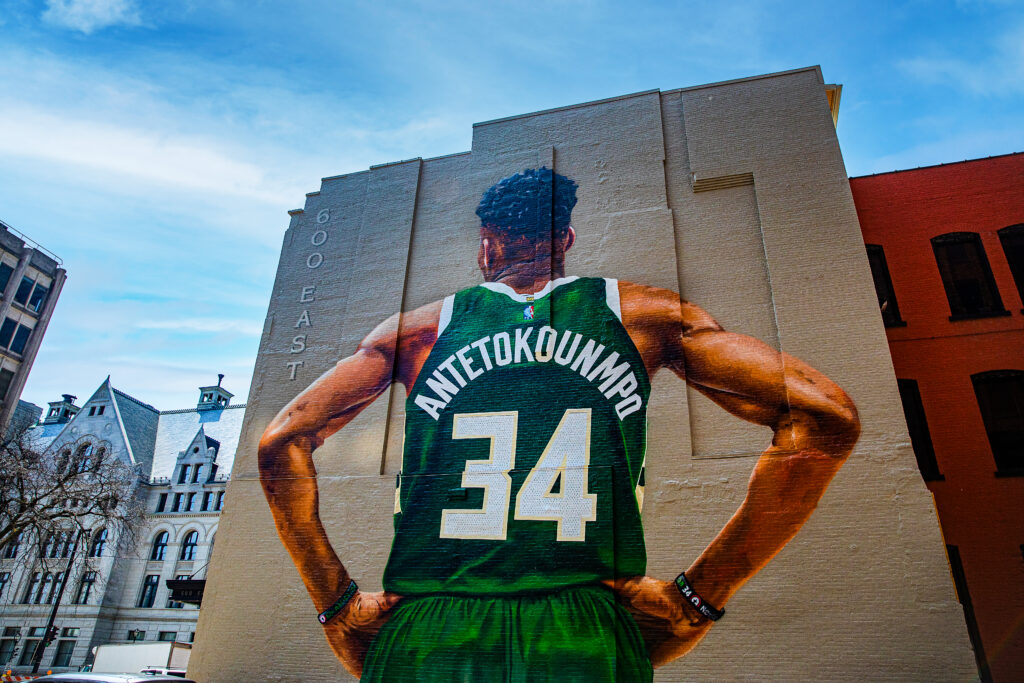 A massive mural of Milwaukee Bucks star Giannis Antetokounmpo covers the side of a downtown building, depicting the player from behind in a green No. 34 jersey with “600 East” painted vertically beside him.