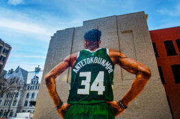 A massive mural of Milwaukee Bucks star Giannis Antetokounmpo covers the side of a downtown building, depicting the player from behind in a green No. 34 jersey with “600 East” painted vertically beside him.