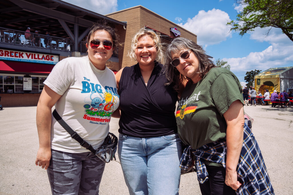 Three women pose together on the Summerfest grounds near the Greek Village food stand, smiling in casual summer clothing under a bright blue sky.