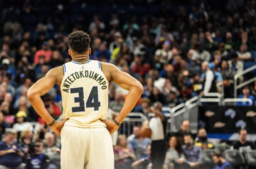 Milwaukee Bucks star Giannis Antetokounmpo stands on the court with his hands on his hips, viewed from behind in a white No. 34 jersey, as a packed arena crowd fills the background.