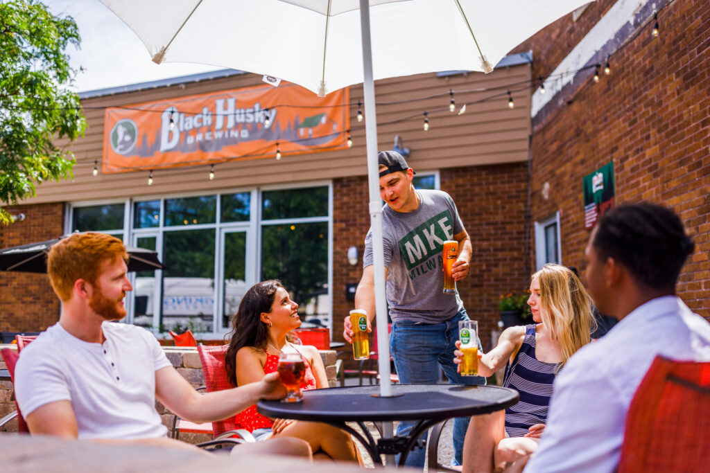 People relax on an outdoor brewery patio beneath a large white umbrella as a server delivers beers to a table, with a sign on a brick building denoting Black Husky Brewing and string lights visible in the background.