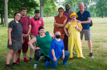 A group of nine people pose together on a grassy park lawn, smiling for a photo, with several wearing brightly colored outfits and one person dressed in yellow overalls and a matching bucket hat.