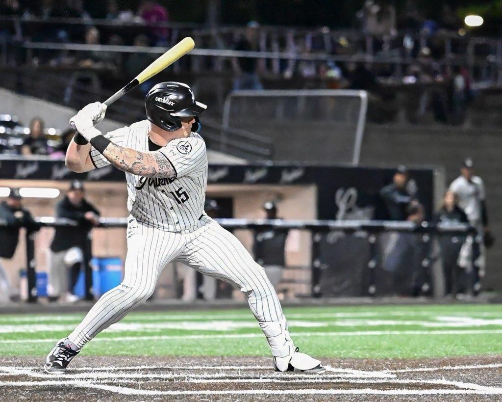 A baseball player in a black pinstriped uniform and black helmet prepares to swing during a nighttime game, with teammates and spectators visible in the background.