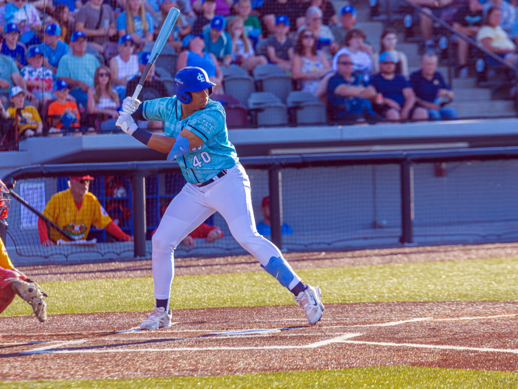 A baseball player in a light blue jersey and white pants waits in the batter’s box with his bat raised, as fans watch from the stands behind home plate.
