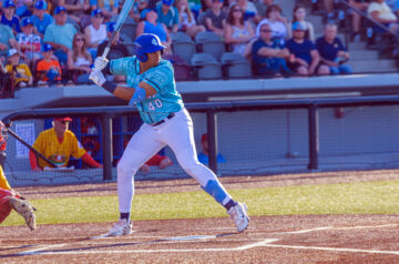 A baseball player in a light blue jersey and white pants waits in the batter’s box with his bat raised, as fans watch from the stands behind home plate.