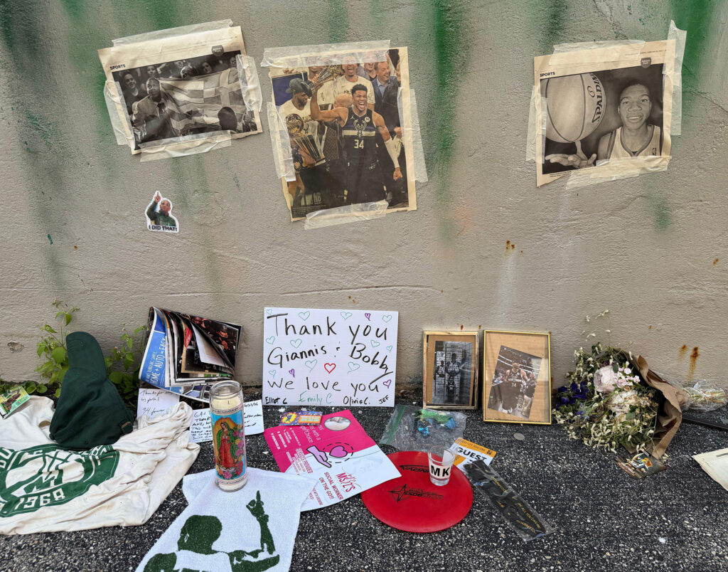 A memorial dedicated to Giannis Antetokounmpo lines the base of a wall, featuring taped newspaper pages, framed photos, flowers, candles, handwritten notes, and Bucks-themed memorabilia, including a sign reading, “Thank you Giannis & Bobby, we love you.”