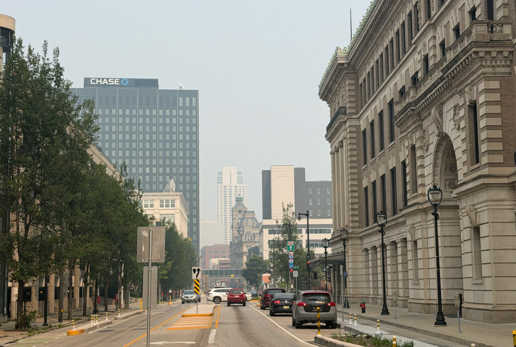 A hazy downtown Milwaukee streetscape shows the Chase Tower and the historic Milwaukee County Courthouse along a city street, with reduced visibility from smoky air softening the skyline in the background.
