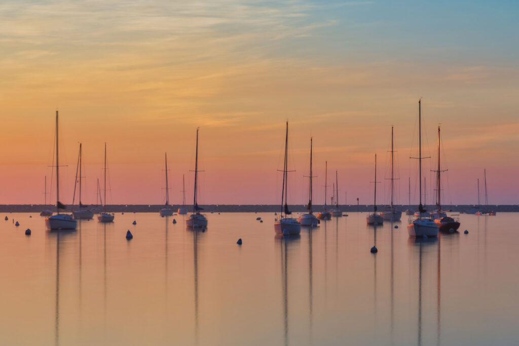 Sailboats sit at anchor on calm water at sunset, their masts reflected on the glassy surface beneath a sky glowing with soft shades of orange, pink, and blue.