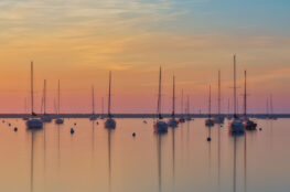 Sailboats sit at anchor on calm water at sunset, their masts reflected on the glassy surface beneath a sky glowing with soft shades of orange, pink, and blue.
