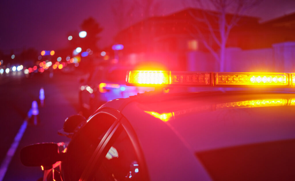 A police vehicle’s flashing red, blue and amber emergency lights illuminate a city street at night, with additional police vehicles and traffic cones blurred in the background.