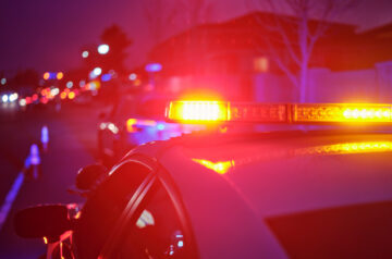 A police vehicle’s flashing red, blue and amber emergency lights illuminate a city street at night, with additional police vehicles and traffic cones blurred in the background.