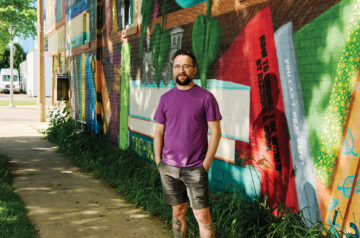 A man with glasses and leg tattoos standing on a sidewalk next to a colorful mural painted he painted on the side of a brick building.