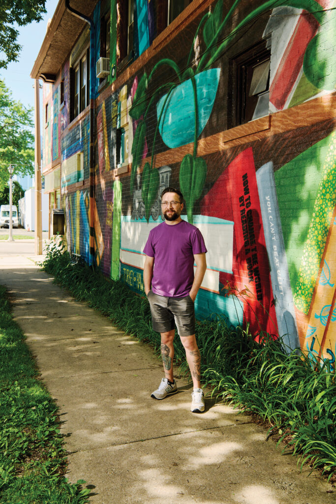 A man with glasses and leg tattoos standing on a sidewalk next to a colorful mural painted he painted on the side of a brick building.