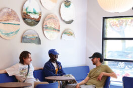 Three people sitting and chatting on a blue cushioned booth in a bright cafe, surrounded by round decorative wall art and modern seating.